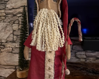 Pode incluir: Boneca artesanal do Pai Natal com túnica e capuz vermelhos, e longa barba branca. A boneca tem um rosto bege com olhos costurados e um bastão de doces. Decoração natalícia festiva.