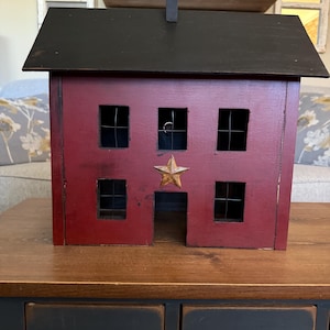 May include: A red wooden house-shaped decorative box with a black roof and chimney. The house has six square windows with wire mesh and a gold star accent. The box sits on a wooden surface.