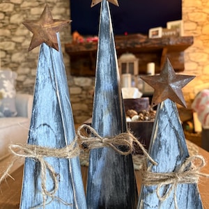 May include: Three rustic wooden Christmas trees with star toppers. The trees are painted in shades of grey and black, with a distressed finish. Each tree is wrapped with twine. The stars are made of rusty metal.