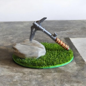 May include: A miniature silver ice axe with a brown leather wrap on the handle is displayed on a small green grass base with a gray rock.