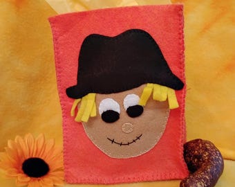 May include: An orange felt gift bag with a stitched scarecrow face. The scarecrow has a black hat, yellow hair, white eyes, and a brown face.