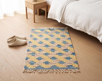 May include: A rectangular area rug featuring a repeating geometric pattern in shades of yellow, blue, and cream. The rug has a textured, woven surface and is finished with short, cream-colored fringe along the top and bottom edges.