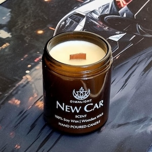 May include: A brown glass jar candle with a wooden wick. The candle is labeled "New Car Scent" and is made with 100% soy wax. The candle has a cream-colored wax and a wooden wick. The candle is hand-poured.