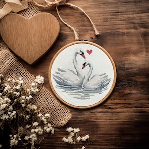 May include: A cross-stitch embroidery of two swans with a red heart above them, set in a wooden hoop. A wooden heart and dried flowers are arranged on a wooden surface with burlap and twine.