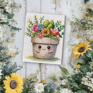 May include: Cross-stitch art of a smiling flower pot filled with colorful flowers. The pot is beige with a smiling face. The flowers are pink, yellow, blue, and green. The artwork is on a white background with floral accents.