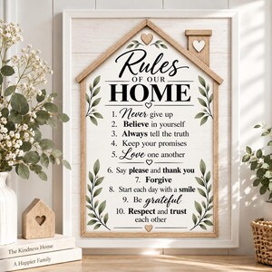 May include: A house-shaped wooden wall sign featuring the text Rules of Our Home and a list of ten numbered rules. The sign has a white background with green leaf illustrations, small heart accents, and a light brown wooden frame and chimney.