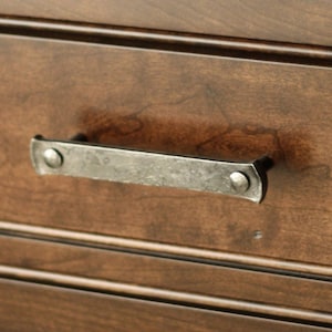 May include: A silver metal drawer pull with a hammered finish and rounded edges. The pull has two visible rivets on each end.