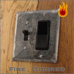 May include: A textured, gray metal light switch plate with two black switches, mounted on a wooden surface. The words "FIRE COOKED" are at the bottom of the image, with a flame graphic in the upper right corner.
