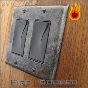 May include: A double rocker light switch plate with a dark, hammered metal finish and a textured, uneven surface. The plate features two matte black rectangular switches and is secured by four visible screws. The text FIRE COOKED is present.