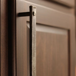 May include: A silver metal cabinet door pull with a hammered finish. The pull is attached to a dark brown wooden cabinet door.