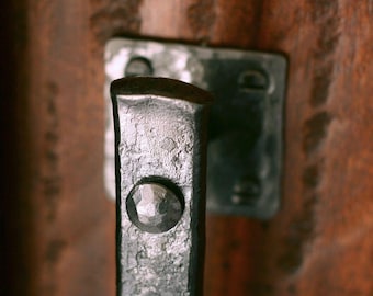 May include: A dark gray, hand-forged metal door handle with a textured, hammered finish. The handle features a rectangular mounting plate secured with visible rivets, showcasing a rugged, industrial appearance with a distinct metallic sheen.