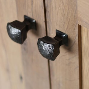May include: Close-up of two black, textured cabinet knobs mounted on light brown wooden cabinet doors. The knobs have a rustic, hammered metal appearance, adding a touch of industrial design to the furniture.