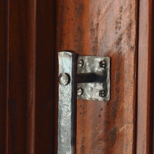 May include: Close-up of a rustic, forged metal door handle and plate, mounted on a dark wooden door. The handle is a vertical bar with a rounded top, and the plate is square with decorative rivets.