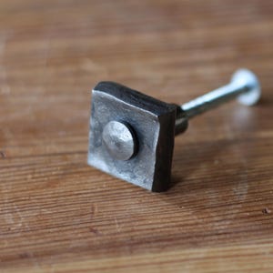 May include: A square, dark metal cabinet knob with a circular accent in the center. The knob is attached to a silver-colored screw. The knob is set against a wooden surface.