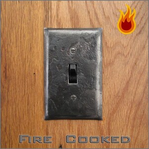 May include: A rectangular, dark gray, hammered metal light switch plate with a central black toggle switch labeled ON. The plate is secured with two matching screws. A stylized orange and yellow flame icon appears in the top right corner.