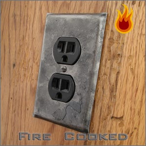 May include: A weathered, gray metal electrical outlet cover with two black outlets. The cover is mounted on a wooden surface. The words "FIRE COOKED" are at the bottom of the image.