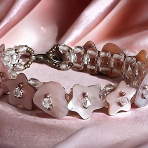 Puede incluir: Un brazalete de cuentas rosa y blanco con un cierre plateado. Las cuentas tienen forma de estrellas y corazones.