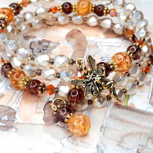 Könnte beinhalten: Ein dreireihiges Armband mit schillernden weißen Perlen, orangefarbenen und braunen Perlen und einem goldenen Feen-Anhänger. Das Armband hat einen baumelnden Anhänger aus orangefarbenen und braunen Perlen.
