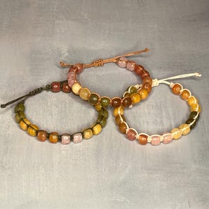 May include: Three beaded bracelets in shades of yellow, orange, pink, and green. The bracelets are made with small, round beads and knotted cords. The bracelets are displayed on a gray surface.