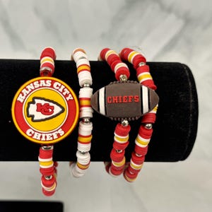 May include: A set of beaded bracelets in red, yellow, white, and silver. One bracelet features a Kansas City Chiefs logo, and another has a football charm with the word "CHIEFS". The bracelets are displayed on a black surface.