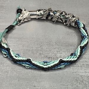 May include: Braided bracelet with a dragon head clasp and silver-toned accents. The bracelet features a mix of black, blue, and teal threads. The bracelet is approximately 7 inches in circumference.