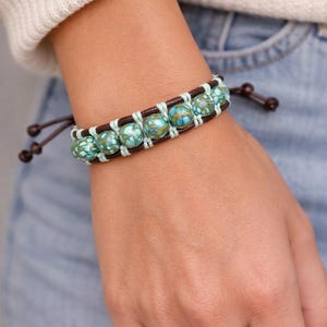 May include: A beaded bracelet with turquoise and brown accents. The bracelet features round, patterned beads in shades of turquoise and brown, strung together with light blue cord and dark brown leather. The bracelet is worn on a wrist.