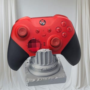 May include: A red Xbox controller with black grips sits on a grey, textured stand. The controller has a central Xbox button, red buttons, and black grips. The stand has a classical column design.