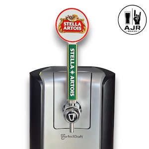 May include: A Stella Artois beer tap handle featuring a round white sign with red trim and the Stella Artois logo, attached to a green rectangular stem with white text. The handle is mounted on a silver and black PerfectDraft beer dispenser.