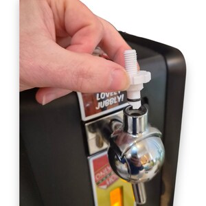 May include: A white plastic threaded tap component with a black rubber O-ring being inserted into a chrome-finished beverage dispenser faucet. The dispenser features a black housing with a label displaying the text LOVELY JUBBLY.