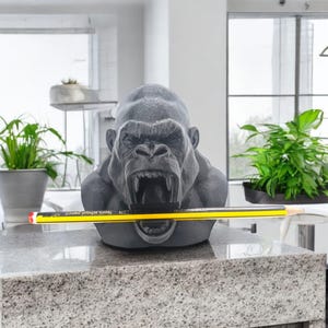 May include: A grey gorilla head sculpture with an open mouth holding a yellow and black pencil. The pencil has the text "Noris school pencil" printed on it. The sculpture is on a granite countertop.