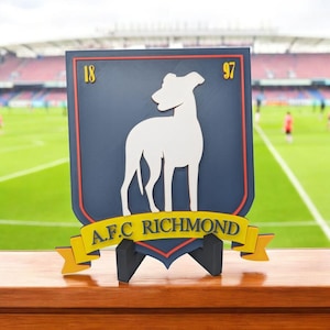 May include: A navy blue and red shield-shaped sign featuring a white dog silhouette, the numbers "18" and "97", and the text "A.F.C RICHMOND" in yellow. The sign is displayed on a black stand, with a football pitch in the background.
