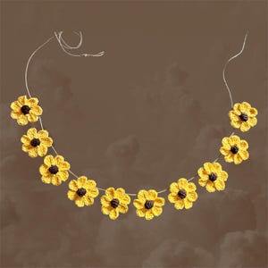May include: A garland of crocheted yellow sunflowers with brown centers, strung on a white twine. The garland is hanging against a brown background.