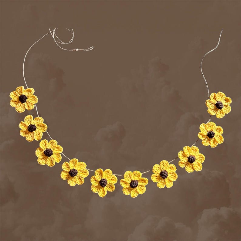 May include: A garland of crocheted yellow sunflowers with brown centers, strung on a white twine. The garland is hanging against a brown background.