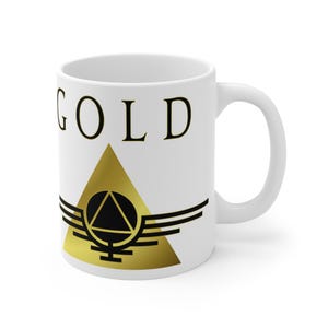 May include: White ceramic mug with the word "GOLD" in black capital letters. A gold triangle with a black circle and wing design is centered below the word. The mug has a white handle.