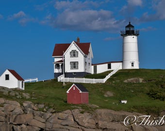 May include: A white lighthouse with a red roof stands on a rocky coastline. A smaller white house with a red roof is to the left of the lighthouse. The lighthouse is surrounded by green grass and trees.