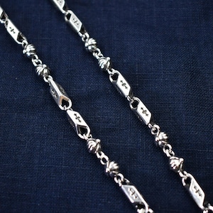 Pode incluir: Close-up de dois colares de prata. Os colares apresentam um padrão repetitivo de elos em forma de diamante com desenhos de cruzes e pequenas contas redondas. O fundo é um tecido azul escuro.