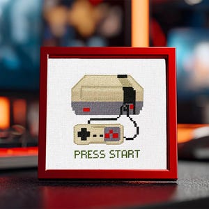 May include: Cross-stitch artwork of a retro video game console and controller, framed in a red square. The console is beige and gray, with a black controller. The text "PRESS START" is below the console in green.