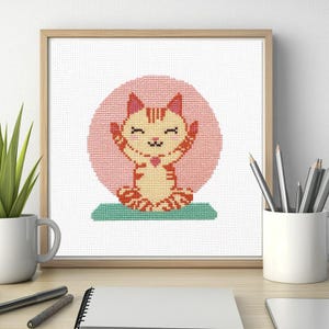 May include: Cross-stitch art of a cartoon cat in a yoga pose. The cat is orange and yellow with a pink heart and is set against a pink circle. The cat is on a green mat, framed in a light wood frame.