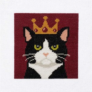 May include: Cross-stitch portrait of a black and white cat wearing a gold crown with red jewels. The cat has green eyes and a pink nose, set against a burgundy background. The artwork is on a white fabric.
