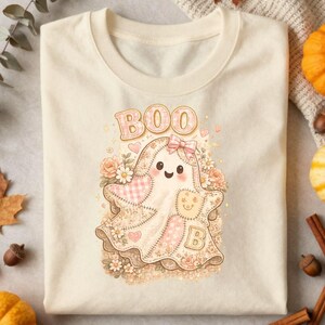 Könnte beinhalten: Ein cremefarbenes T-Shirt mit einer Grafik eines niedlichen Geistes, der eine rosa Schleife trägt. Der Geist ist mit Patchwork-Mustern, Blumen und Herzen verziert. Der Text BOO ist in hellrosa Schrift über dem Geist gedruckt, und der Buchstabe B ist unten zu sehen.
