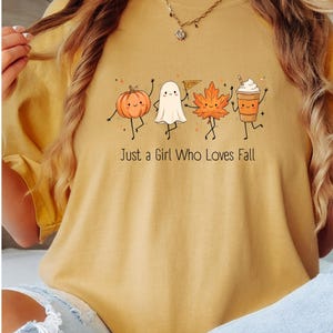 Könnte beinhalten: Ein senfgelbes T-Shirt mit einem Grafikdruck von tanzendem Kürbis, Geist, Ahornblatt und Kaffeebecher. Der Text "Just a Girl Who Loves Fall" ist in einer einfachen schwarzen Schrift unter den Figuren aufgedruckt. Das Shirt hat einen Rundhalsausschnitt.