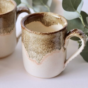 May include: Two ceramic mugs with brown and white glaze. The mugs have a rounded shape and a handle. The mugs are sitting on a white surface.