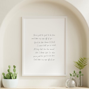Può includere: Una stampa incorniciata con il testo "You're just too good to be true" in elegante scrittura corsiva. La cornice bianca circonda il testo, creando un'estetica minimalista. La stampa è esposta su una mensola con piante decorative.
