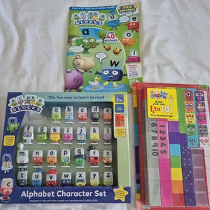 May include: Three educational toy sets for children. The sets include an alphabet character set with figurines, a book with stickers, and a number blocks set. The packaging features colourful illustrations and the text "Alphabet Character Set" and "Number Blocks".