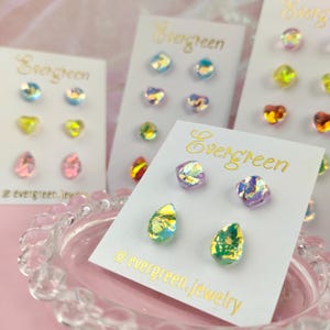 May include: Assortment of iridescent glass earrings in various shapes, including teardrops, squares, and hearts. The earrings are displayed on white cards with the text "Evergreen" printed in gold. The cards are on a white surface.