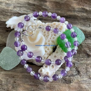 Può includere: Un braccialetto con perline alternate di ametista viola e quarzo trasparente. Il braccialetto è assicurato con una chiusura argentata. Il testo "Amethyst and Clear Quartz" è visibile. Il braccialetto è esposto su una superficie di legno.