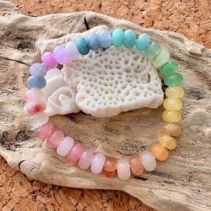 Peut inclure: Un bracelet de perles colorées aux teintes arc-en-ciel, allant du rose à l'orange, en passant par le jaune, le vert, le bleu et le violet. Les perles ovales sont enfilées, posées sur du bois flotté et une structure blanche ressemblant à du corail.