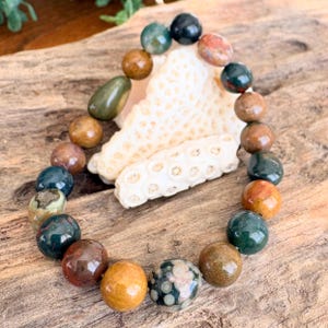 Puede incluir: Un brazalete de cuentas con una variedad de piedras preciosas pulidas de colores, incluyendo verde, marrón y patrones moteados. Las cuentas redondas se exhiben sobre un trozo de madera flotante.