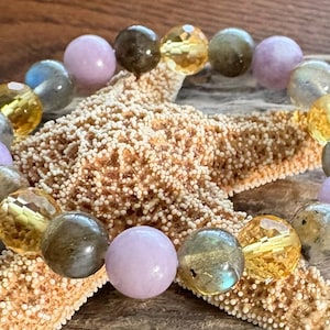 May include: A beaded bracelet featuring a variety of gemstone beads in shades of lavender, gold, and gray. The bracelet is resting on a starfish, creating a beachy aesthetic. The beads are approximately 1 cm in diameter.
