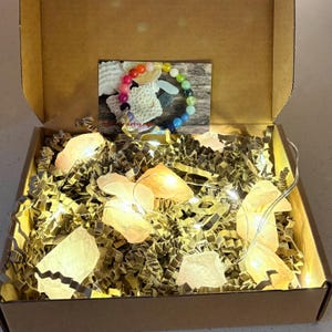 Könnte beinhalten: Ein Karton gefüllt mit goldenem Zerkleinerungspapier, beleuchtet von kleinen Lichterketten und mehreren großen, leuchtenden, hellrosa Kristallen. Eine kleine Karte mit einem bunten Perlenarmband ist sichtbar.